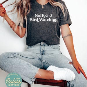 May include: A dark gray t-shirt with the text "Coffee & Bird Watching" in white. The model is wearing the shirt with blue jeans and white sneakers.