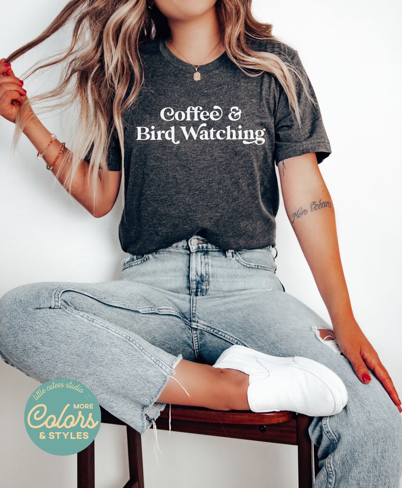 May include: A dark gray t-shirt with the text "Coffee & Bird Watching" in white. The model is wearing the shirt with blue jeans and white sneakers.