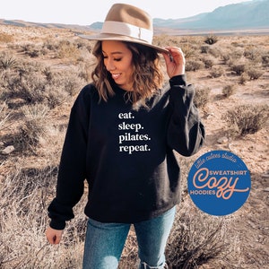 May include: A black sweatshirt with the text "eat. sleep. pilates. repeat." in white. The sweatshirt is being worn by a person in a desert setting. The image also includes a logo for "little cutes studio" with the text "SWEATSHIRT Cozy HOODIES".