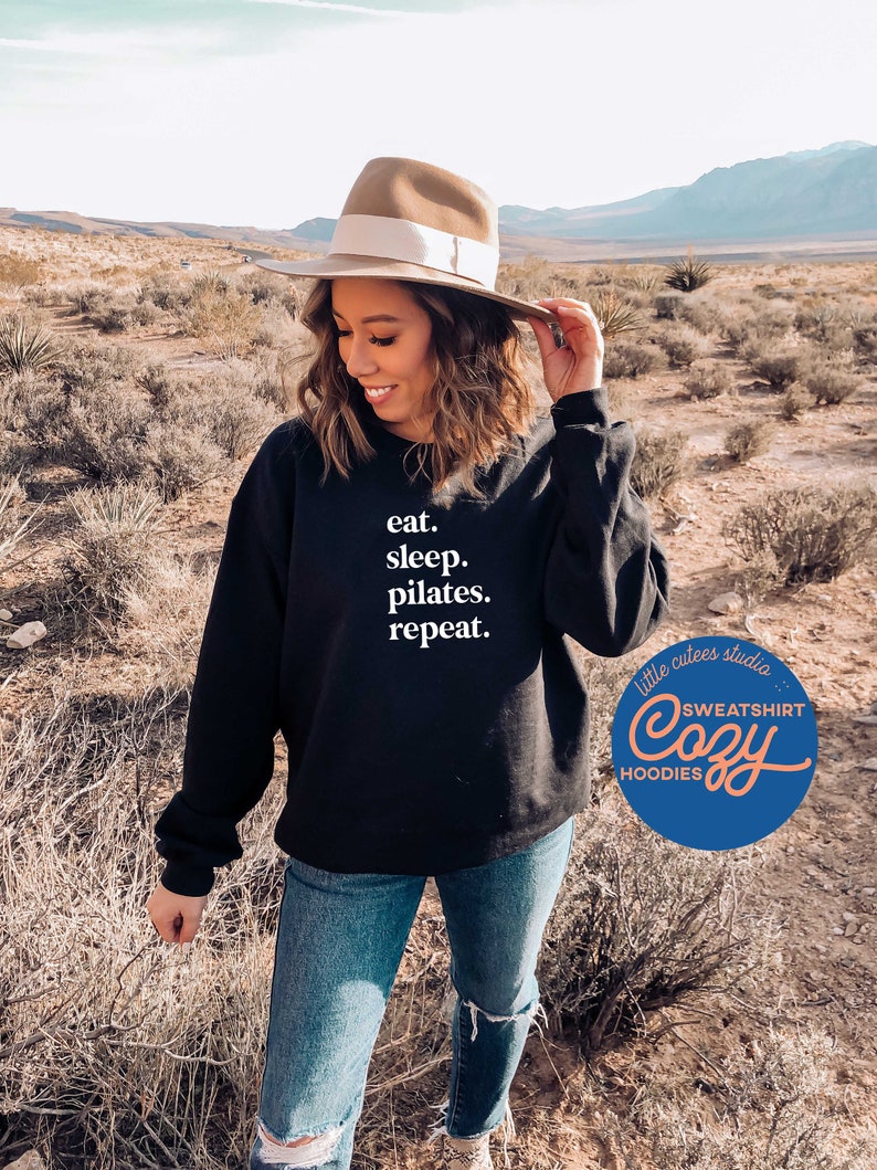 May include: A black sweatshirt with the text "eat. sleep. pilates. repeat." in white. The sweatshirt is being worn by a person in a desert setting. The image also includes a logo for "little cutes studio" with the text "SWEATSHIRT Cozy HOODIES".