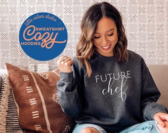 May include: A dark gray sweatshirt with the text "FUTURE chef" in white script. The model is wearing blue jeans and sitting on a couch with a brown and white patterned pillow.