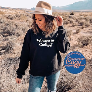May include: A black sweatshirt with the text "Women in Coding" in white lettering. The sweatshirt is being worn by a person in a desert setting. A blue circle with the text "little cutes studio" and "SWEATSHIRT Cozy HOODIES" in white lettering is in the lower right corner of the image.