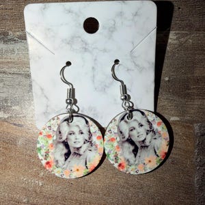 May include: A pair of round dangle earrings featuring a black and white portrait of a blonde woman with voluminous hair, surrounded by a colorful floral border of orange, pink, and green flowers, attached to silver-toned metal earring hooks.
