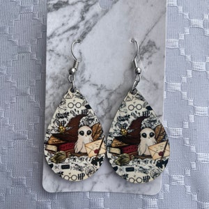 May include: A pair of Harry Potter themed dangle earrings. The earrings are made of wood and feature a white owl, a wizard's hat, and the words "Transfiguration" and "Potions".