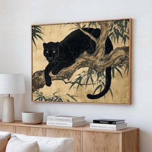May include: A framed artwork depicts a black panther resting on a tree branch against a gold background. The panther's fur is detailed, and its yellow eyes are visible. The artwork is framed in a light-colored wood.