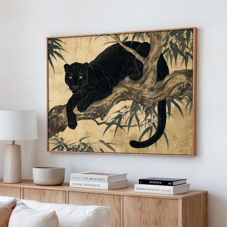 May include: A framed artwork depicts a black panther resting on a tree branch against a gold background. The panther's fur is detailed, and its yellow eyes are visible. The artwork is framed in a light-colored wood.