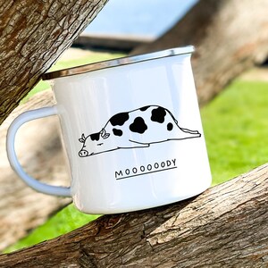 May include: White enamel camping mug with a black and white illustration of a cow lying down and the text "MOOOOODY".