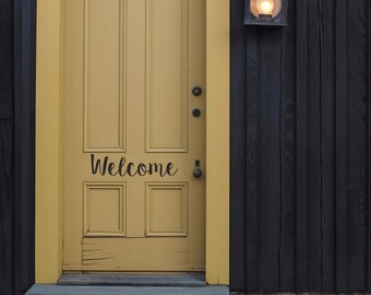 May include: A yellow wooden door with a black welcome sign on a black wooden building with a light fixture on the side.