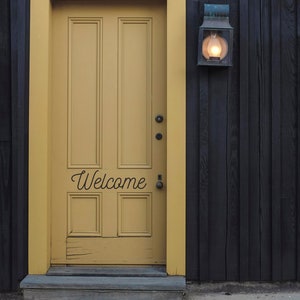 May include: A yellow door with a welcome sign on a black wooden building. A light fixture is mounted on the wall next to the door.