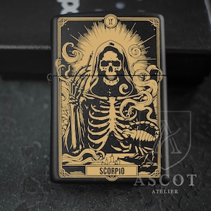 May include: A matte black metal lighter featuring a gold-toned graphic of a hooded skeleton holding a scorpion. The design includes a sunburst halo, swirling smoke, and candles. The word SCORPIO is printed in a rectangular frame at the bottom.