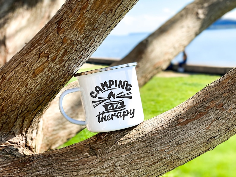 May include: White enamel camping mug with black text that reads "Camping is my therapy". The mug is sitting on a tree branch.