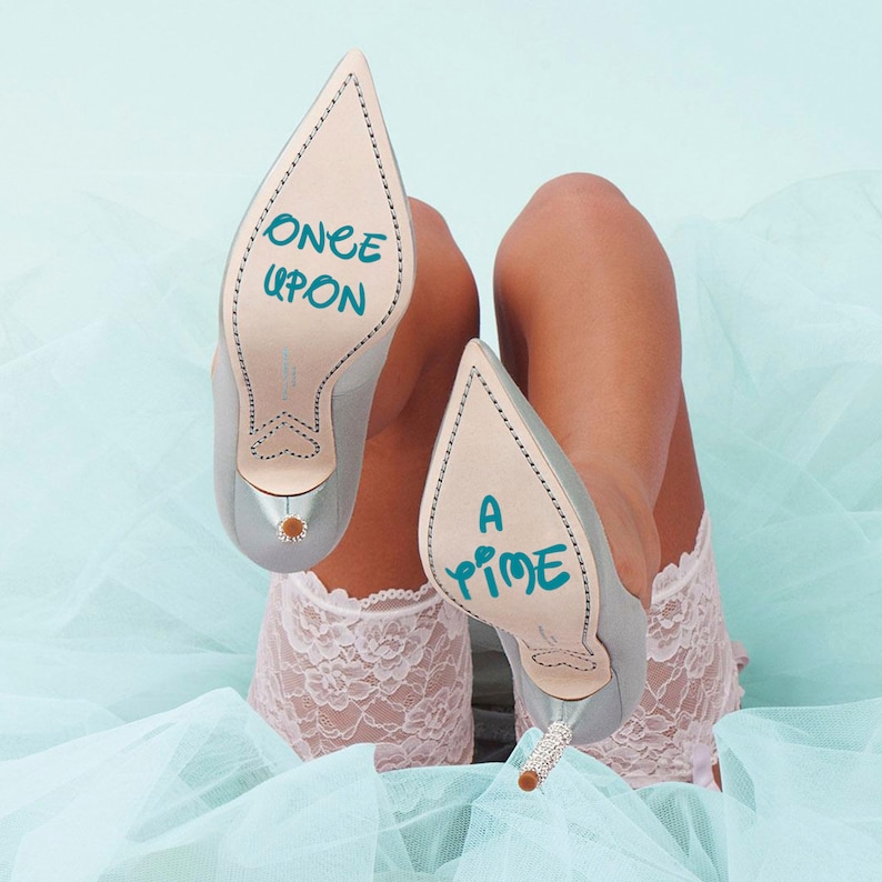 May include: A pair of light blue satin high heels with a pointed toe. The heels have a white lace trim and the soles are inscribed with the words "Once Upon A Time" in blue.