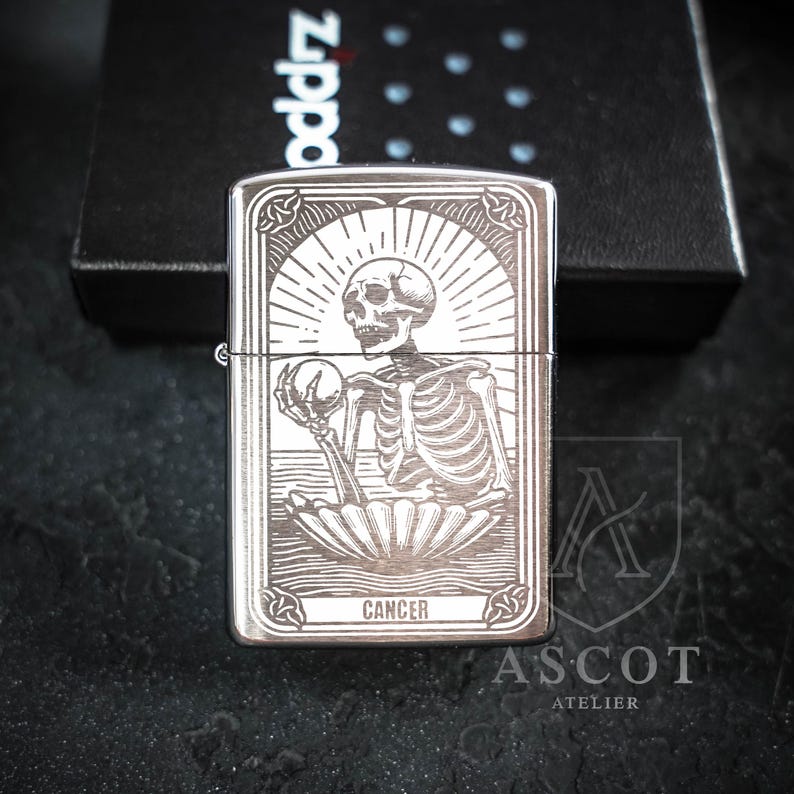 May include: A silver-toned metal lighter featuring an engraved illustration of a skeleton emerging from a shell, holding a sphere. The design includes sunburst rays and the word CANCER at the bottom. The lighter is positioned in front of a black box.