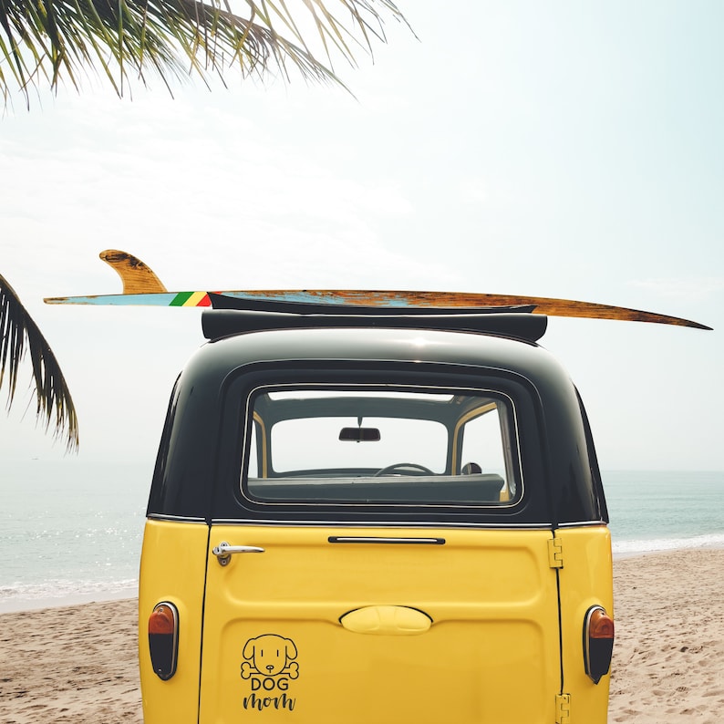 May include: A yellow vintage car with a surfboard on the roof. The car has a black roof and a sticker on the back that says "Dog Mom" with a cartoon dog illustration.