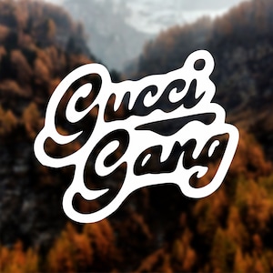 May include: White sticker with black text that reads "Gucci Gang" in a stylized font. The sticker is placed on a blurred background of a forest.