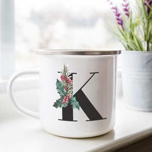 Puede incluir: Taza de esmalte blanco con una letra K negra y una rama de acebo verde con bayas rojas. La taza tiene un borde y un asa plateados.