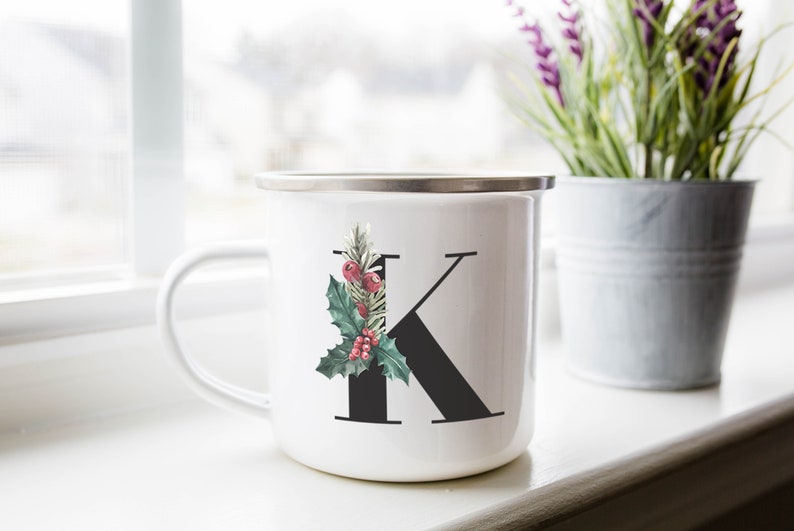 Puede incluir: Taza de esmalte blanco con una letra K negra y una rama de acebo verde con bayas rojas. La taza tiene un borde y un asa plateados.