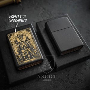 May include: Two matte black metal lighters, one featuring a gold-toned engraving of a skeleton wearing a crown and sitting on a throne with the text LEO, and the other plain. The text FRONT SIDE ENGRAVING is visible with an arrow pointing to the design.