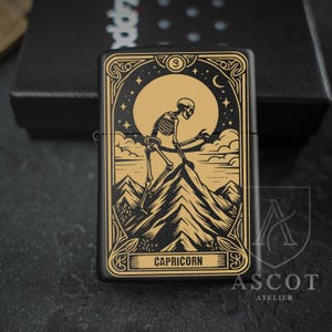 May include: A matte black metal lighter featuring a gold-toned engraved design of a skeleton climbing a mountain peak under a large moon and stars. The rectangular frame includes ornate border details and the word CAPRICORN printed at the bottom.
