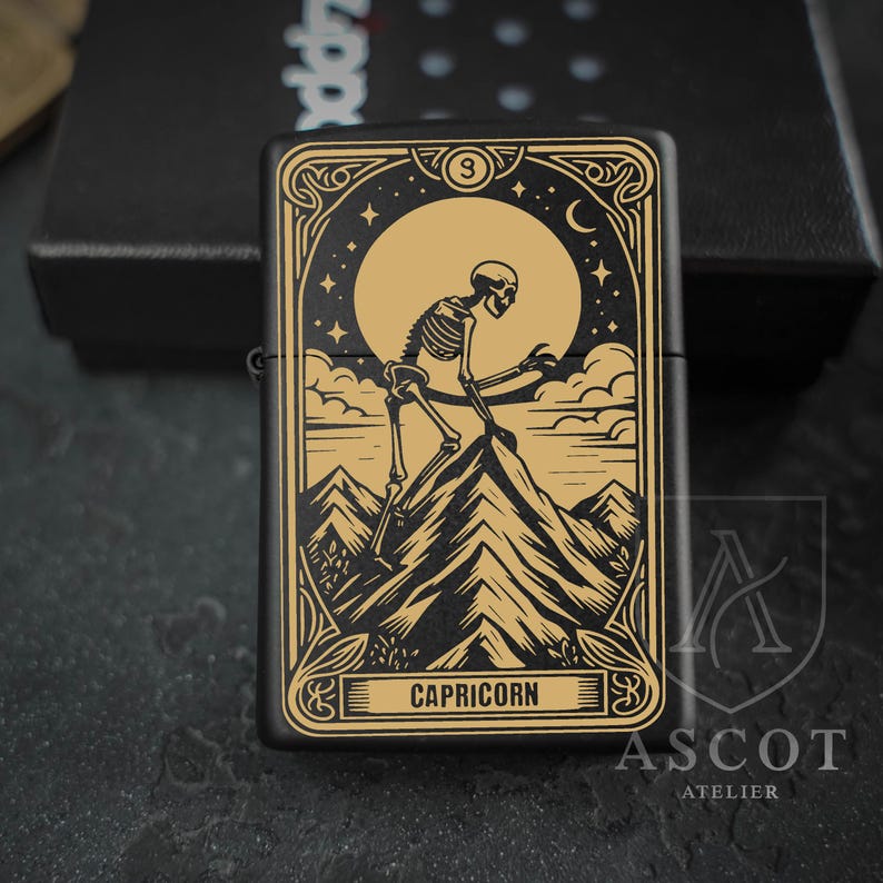 May include: A matte black metal lighter featuring a gold-toned engraved design of a skeleton climbing a mountain peak under a large moon and stars. The rectangular frame includes ornate border details and the word CAPRICORN printed at the bottom.
