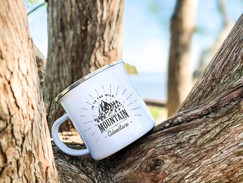 May include: White enamel camping mug with a black and white graphic of a mountain range and the text "Into the Wild. Mountain Adventure".