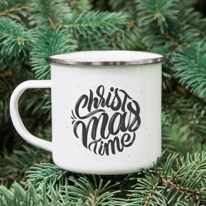 Puede incluir: Taza de esmalte blanca con un dise&ntilde;o en blanco y negro que dice "Christmas time" en una fuente manuscrita. La taza est&aacute; sentada sobre un lecho de agujas de pino verdes.
