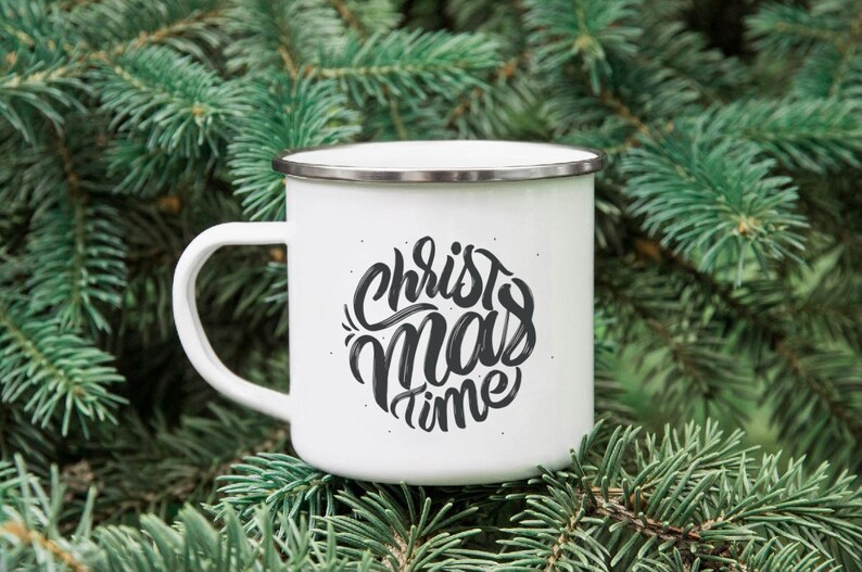 Puede incluir: Taza de esmalte blanca con un dise&ntilde;o en blanco y negro que dice "Christmas time" en una fuente manuscrita. La taza est&aacute; sentada sobre un lecho de agujas de pino verdes.