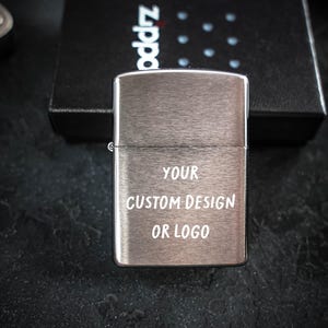 May include: A brushed silver metal lighter with a rectangular shape and a hinged lid. The front surface features the white text YOUR CUSTOM DESIGN OR LOGO engraved in a simple sans-serif font. The lighter is positioned in front of a black box.