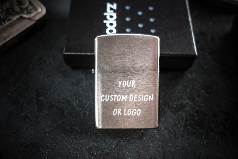 May include: A brushed silver metal lighter with a rectangular shape and a hinged lid. The front surface features the white text YOUR CUSTOM DESIGN OR LOGO engraved in a simple sans-serif font. The lighter is positioned in front of a black box.