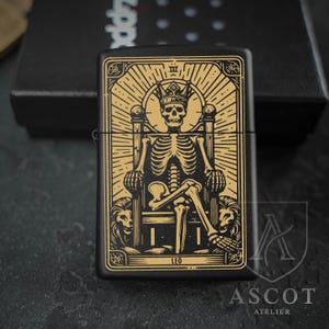 May include: A matte black metal lighter featuring a gold-colored engraved design of a skeleton wearing a crown and sitting on a throne flanked by two lions. The design includes the text LEO at the bottom and Roman numeral III at the top.