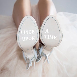 May include: A pair of white high-heeled shoes with butterfly embellishments. The soles of the shoes are white and have the words "Once Upon" and "A Time" written on them.