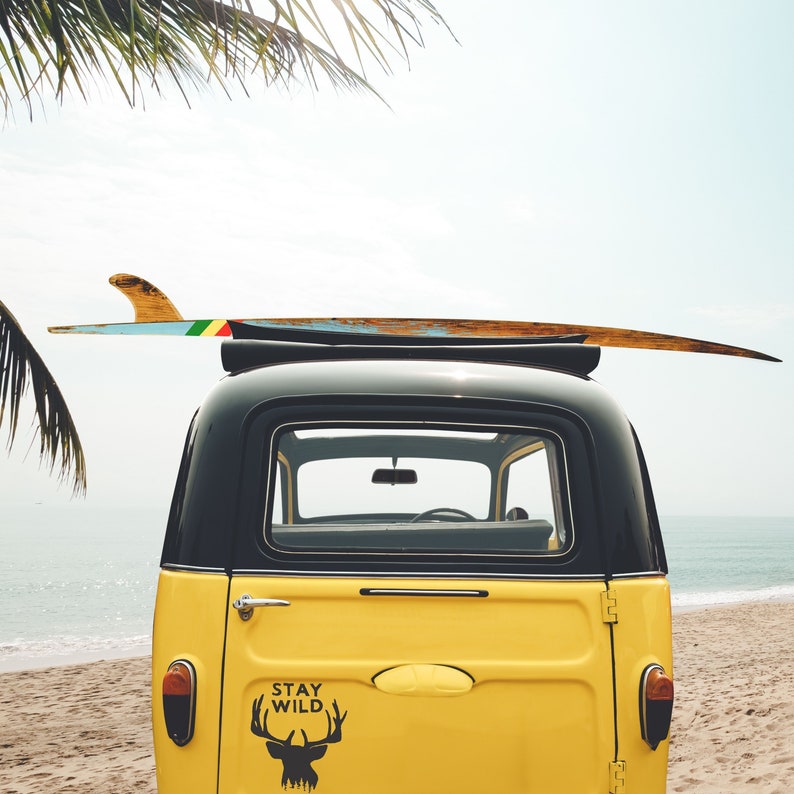 May include: A yellow vintage car with a surfboard on top. The car has a black roof and black trim. The back window has a sticker that says "Stay Wild" with a deer silhouette.