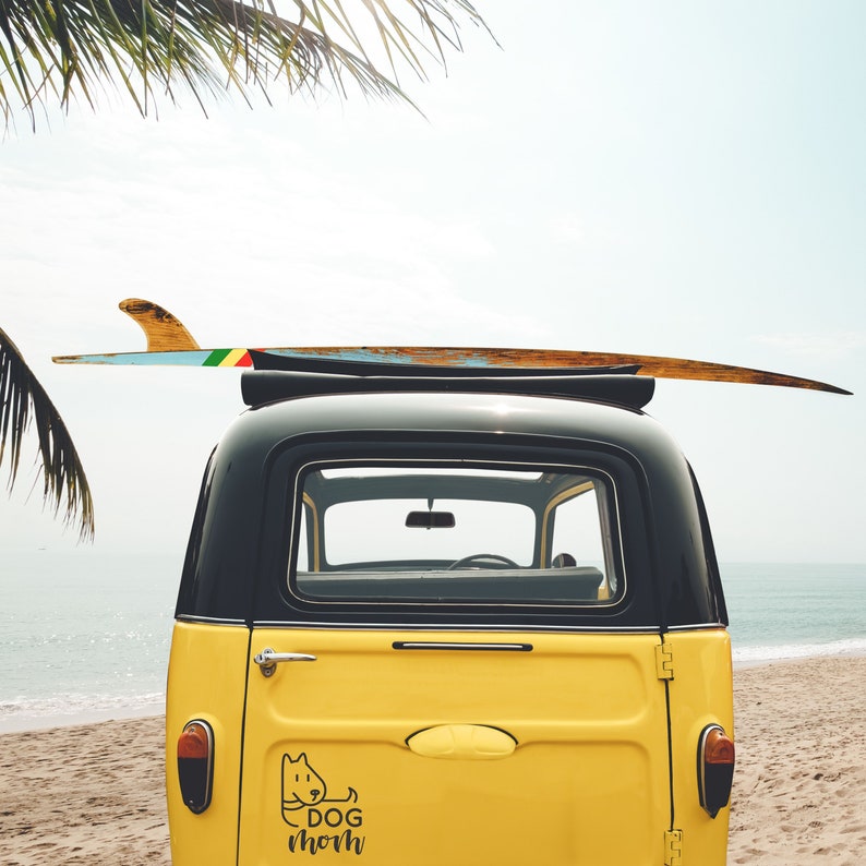 May include: A yellow vintage car with a surfboard on the roof. The car has a black roof and a sticker on the back that says "Dog Mom".