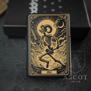 May include: A matte black metal lighter featuring a gold-toned engraved illustration of a skeleton with ram horns holding a sword. The design includes a sun, moon, and clouds, with the text ARIES printed in a rectangular frame at the bottom.