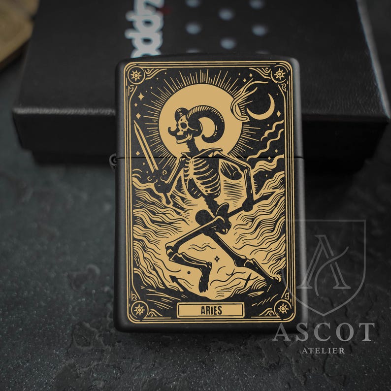 May include: A matte black metal lighter featuring a gold-toned engraved illustration of a skeleton with ram horns holding a sword. The design includes a sun, moon, and clouds, with the text ARIES printed in a rectangular frame at the bottom.