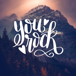 May include: White text on a blurred background of a mountain range with the words "you rock" with a heart symbol on either side of the text.