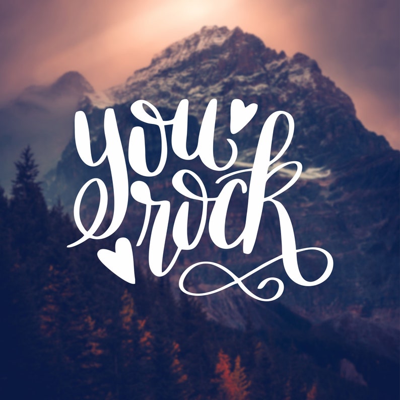 May include: White text on a blurred background of a mountain range with the words "you rock" with a heart symbol on either side of the text.