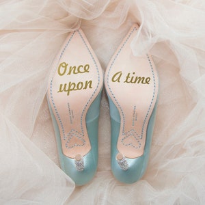 May include: A pair of light blue satin heels with a gold inscription on the soles. The left shoe says "Once upon" and the right shoe says "a time".