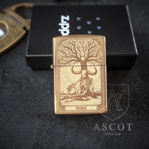 May include: A rectangular brass-toned metal lighter featuring an engraved design of a skeleton with a bull skull head sitting under a tree. The word TAURUS is inscribed at the bottom of the graphic. The lighter is positioned in front of a black box.