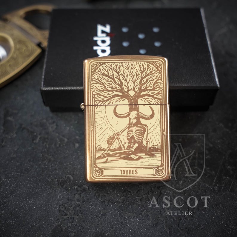 May include: A rectangular brass-toned metal lighter featuring an engraved design of a skeleton with a bull skull head sitting under a tree. The word TAURUS is inscribed at the bottom of the graphic. The lighter is positioned in front of a black box.