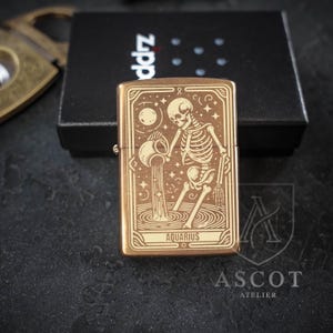 May include: A rectangular brass-toned metal lighter featuring an engraved design of a skeleton pouring water from a pitcher, representing the Aquarius zodiac sign. The word AQUARIUS is printed at the bottom of the design, which is framed by lines.