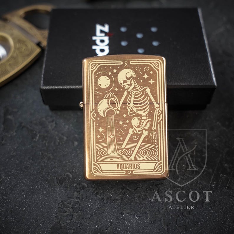 May include: A rectangular brass-toned metal lighter featuring an engraved design of a skeleton pouring water from a pitcher, representing the Aquarius zodiac sign. The word AQUARIUS is printed at the bottom of the design, which is framed by lines.