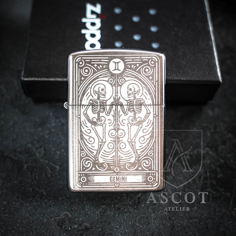 May include: A silver-toned metal lighter featuring a detailed laser-engraved design of two skeletons facing each other, the Gemini zodiac symbol at the top, and the word GEMINI at the bottom, presented in a black Zippo branded box.