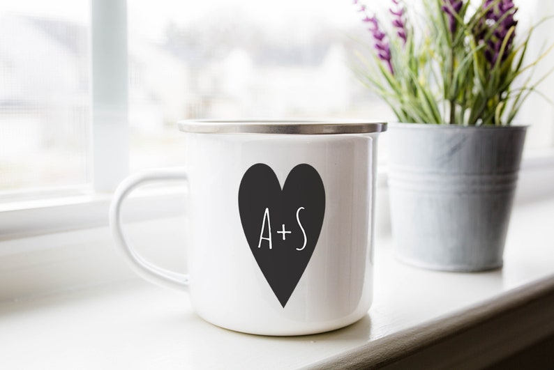 Puede incluir: Taza de esmalte blanco con un coraz&oacute;n negro y el texto "A+S" en ella. La taza tiene un borde plateado y un asa.