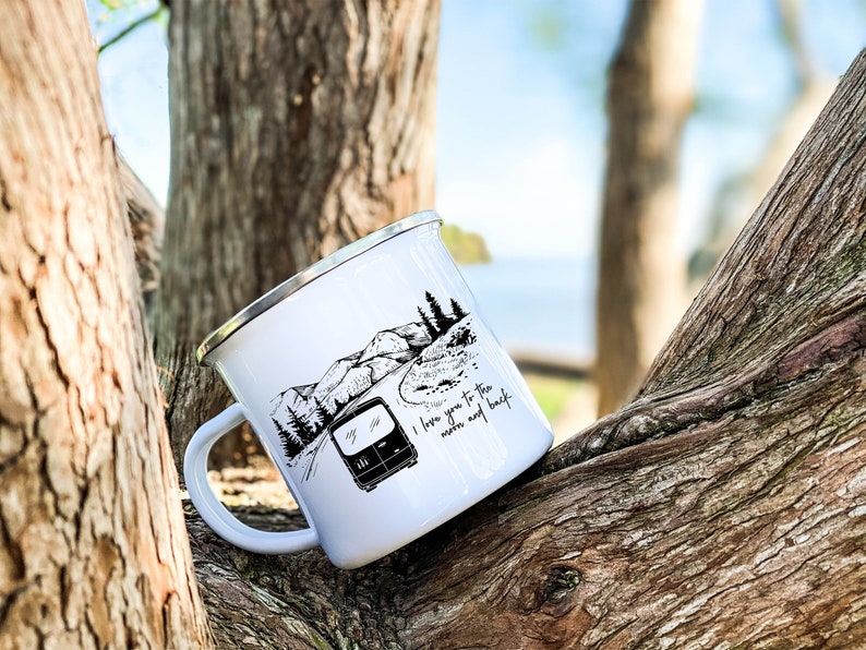 May include: White enamel camping mug with a black and white illustration of a camper van driving on a winding road through mountains. The text "I love you to the moon and back" is printed on the mug.