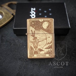 May include: A rectangular bronze-toned metal lighter featuring an engraved tarot card design of a skeleton archer aiming a bow. The image includes a moon, stars, and mountains, with the text SAGITTARIUS at the bottom. The lighter rests on a black box.