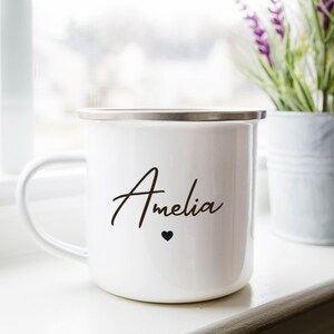 Puede incluir: Taza de esmalte blanco con borde plateado y el nombre "Amelia" escrito en cursiva negra con un peque&ntilde;o coraz&oacute;n negro debajo.