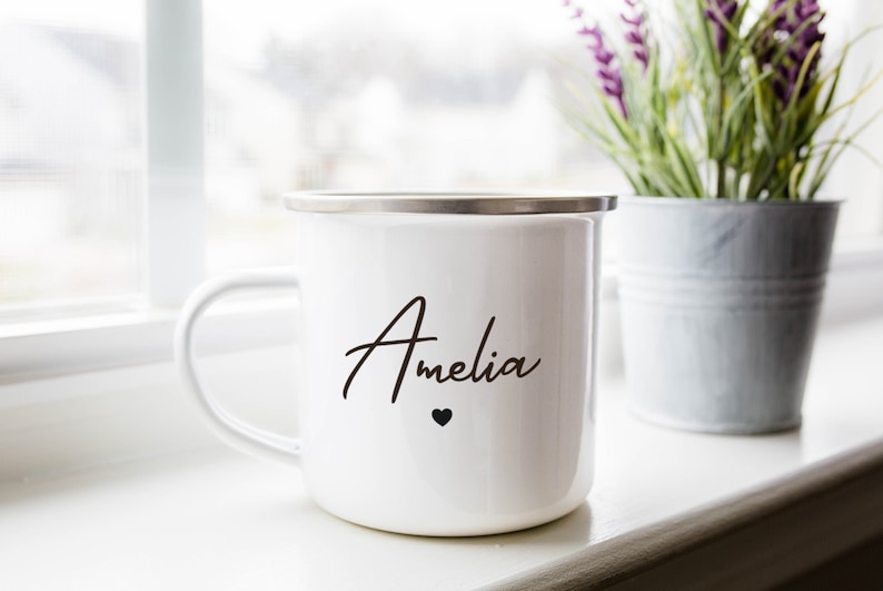 Puede incluir: Taza de esmalte blanco con borde plateado y el nombre "Amelia" escrito en cursiva negra con un peque&ntilde;o coraz&oacute;n negro debajo.