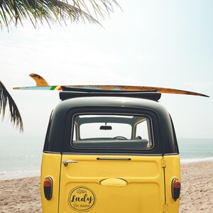 May include: A yellow vintage car with a surfboard on top. The car has a black roof and a sticker on the back that says "Little Lady on board".