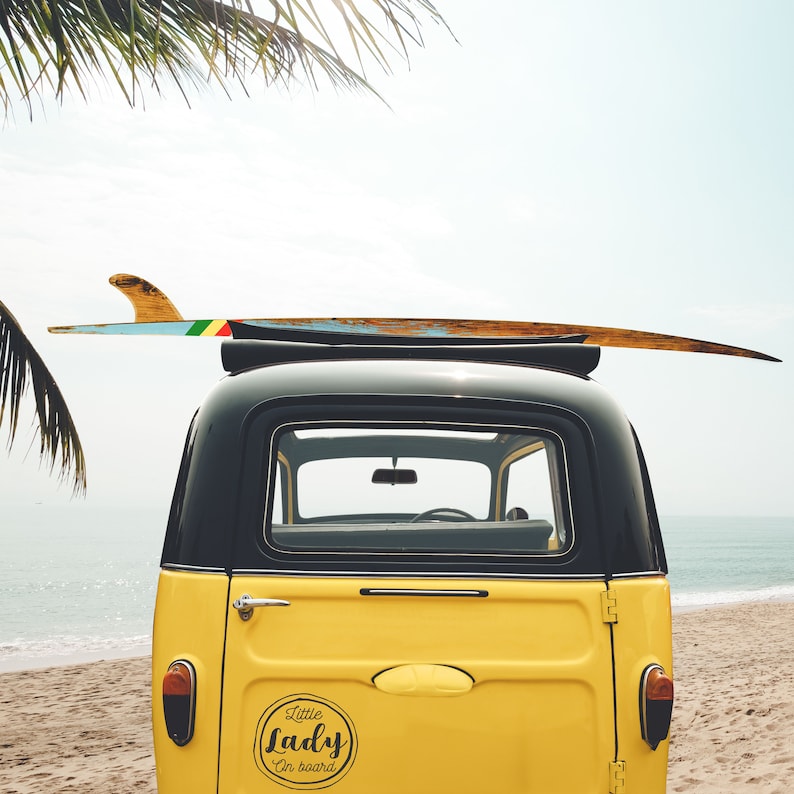 May include: A yellow vintage car with a surfboard on top. The car has a black roof and a sticker on the back that says "Little Lady on board".